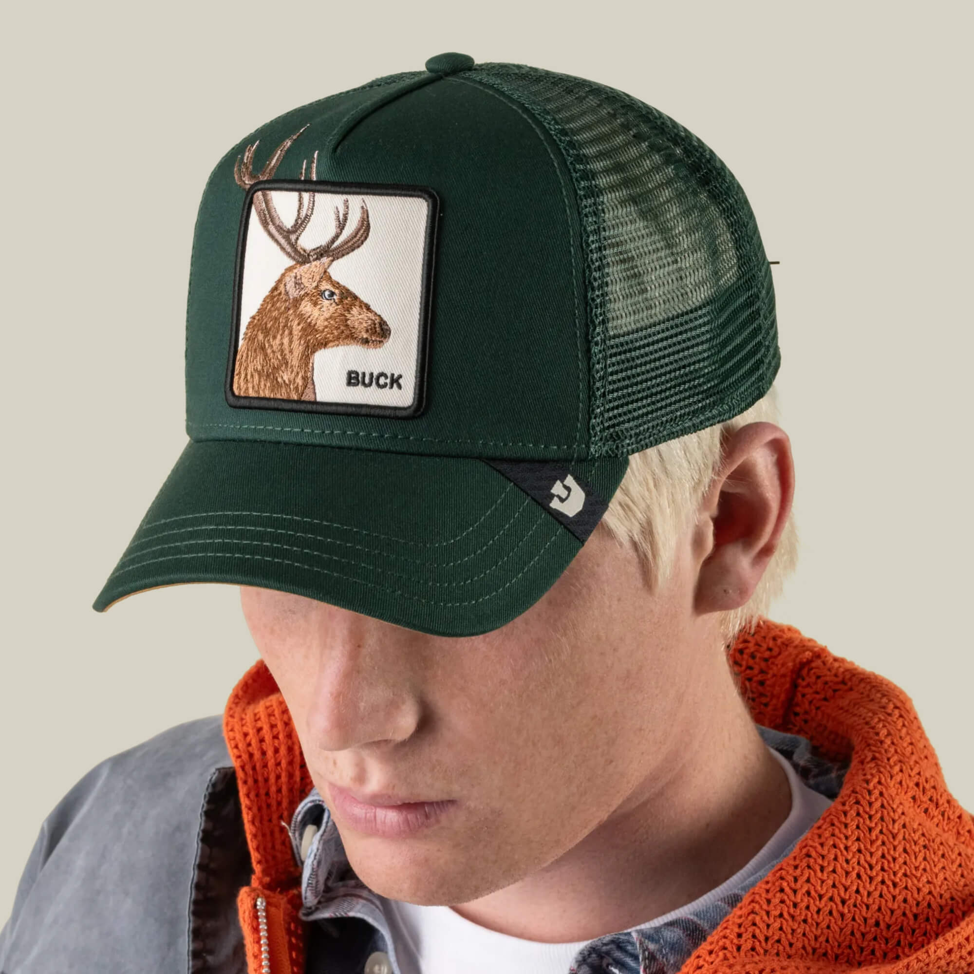 A person wearing Goorin Bros.’ Over the Line Buck green trucker hat—crafted from a breathable mesh polyester-cotton blend and featuring a patch with a brown buck illustration and the word “BUCK”—pairs it with an orange hoodie and gray jacket.