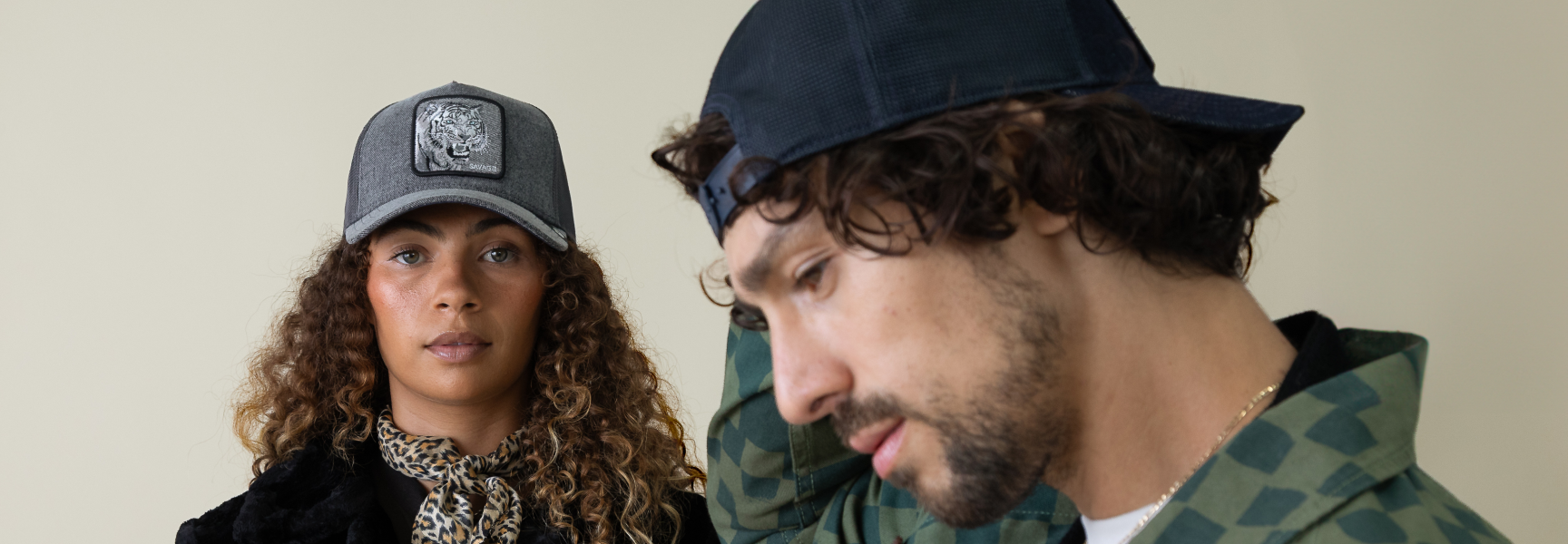 A woman in a gray trucker hat stands in the background, while a man in a dark cap and green patterned shirt is in the foreground, adjusting his hat.