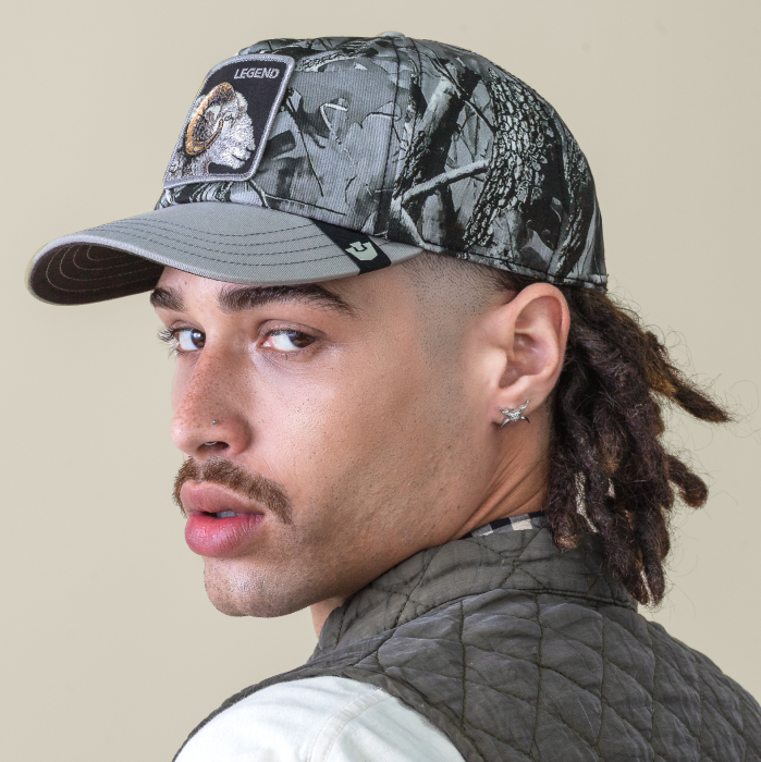 A person with a mustache and pierced ear wears a camo baseball cap featuring bold hat designs and a quilted jacket, looking over their shoulder at the camera.