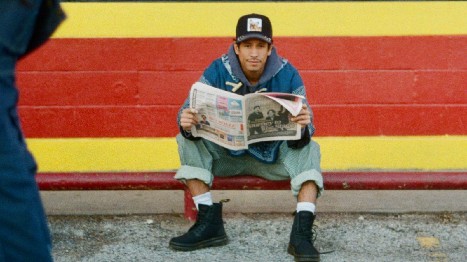 A person wearing a bold hat design and boots sits on a curb in front of a striped red and yellow wall, holding and reading a newspaper.