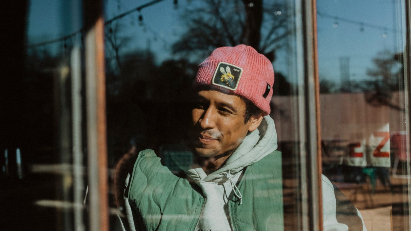 A man wearing a pink beanie with bold hat designs, a green vest, and a gray hoodie stands outside, seen through a glass window with outdoor scenery in the background.
