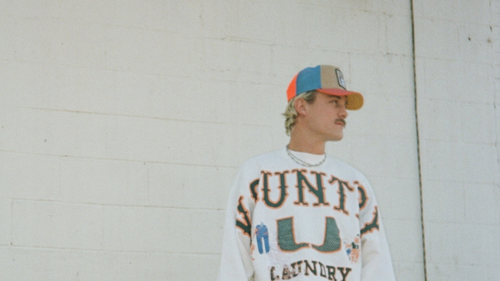 A person wearing a colorful Goorin trucker hat and a white sweatshirt with bold text stands in front of a white brick wall, looking to the side.