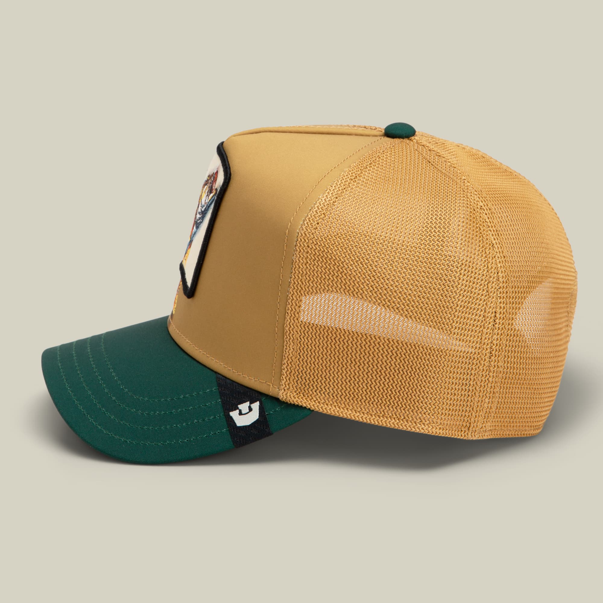 The Goorin Bros. Migration Pro trucker hat features a tan and cream front panel with a mesh back, teal green bill, and an animal patch on the side, displayed against a neutral background.