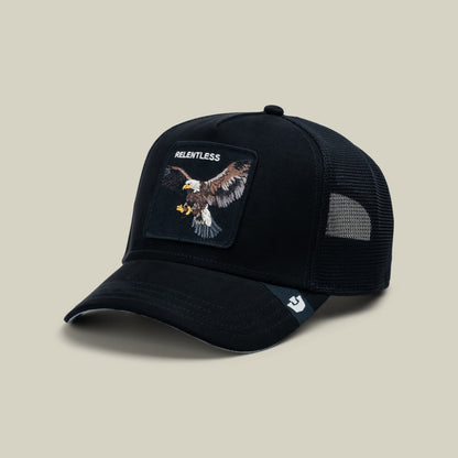 The Goorin Bros. Suede Relentless Eagle Trucker is a black mesh-back cap featuring an embroidered eagle patch with "Relentless" text on the front, displayed on a neutral beige studio backdrop.