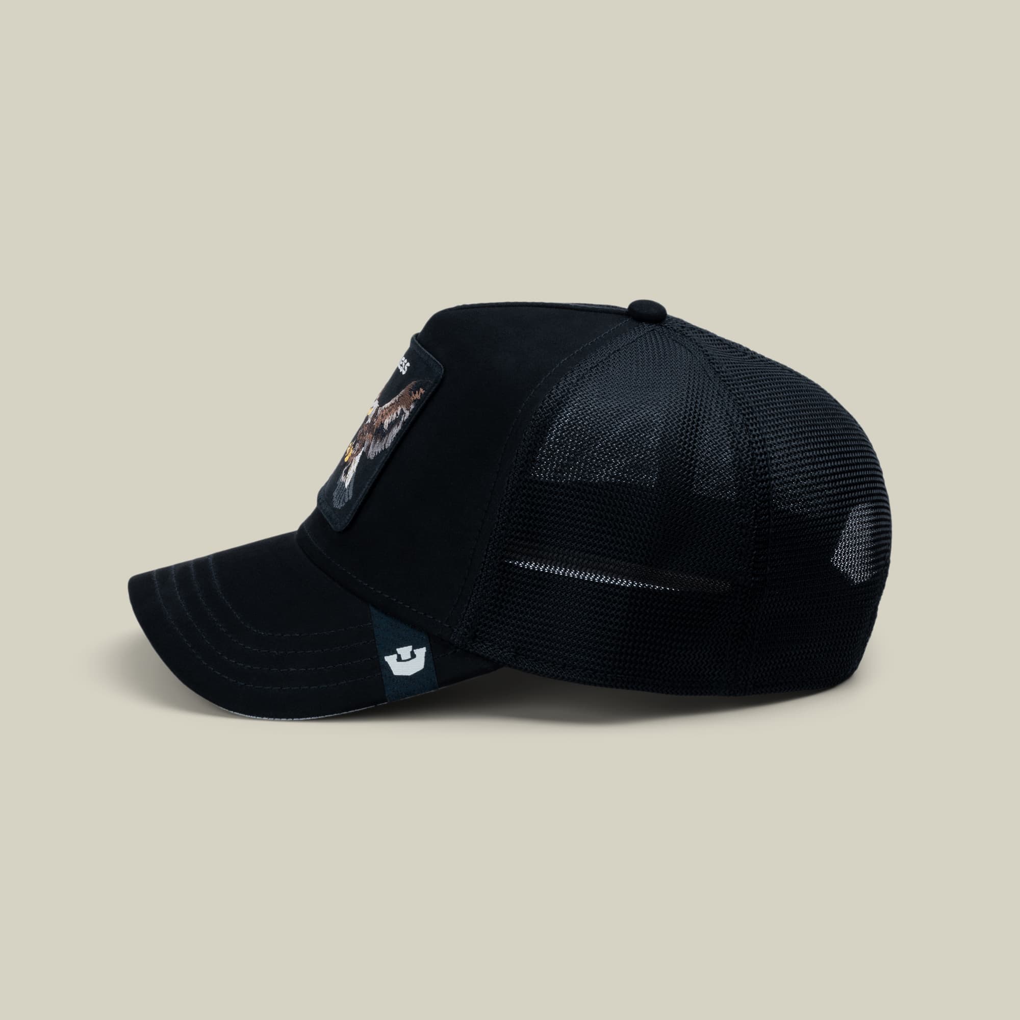 The Goorin Bros. Suede Relentless Eagle Trucker is a black mesh-back cap with suede front panels, featuring an embroidered eagle patch on the side, displayed against a neutral beige background.