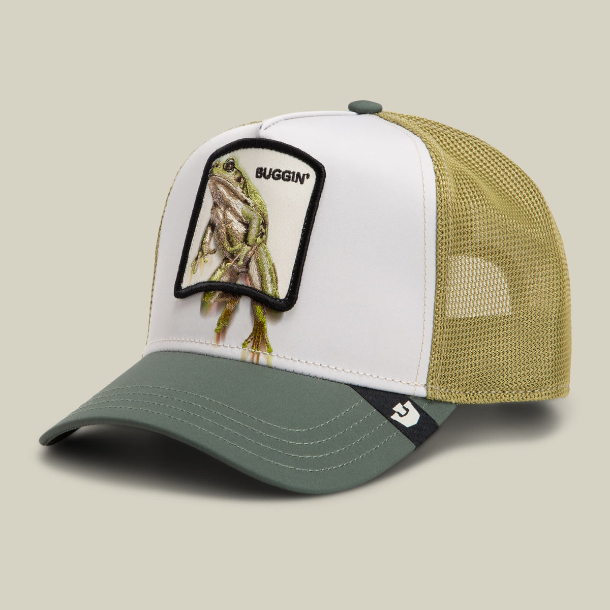 The Goorin Bros. Migration Buggin trucker hat features a white front panel and beige mesh back with a green visor, detailed with an embroidered frog patch and "Buggin'" text. Displayed on a neutral studio background.